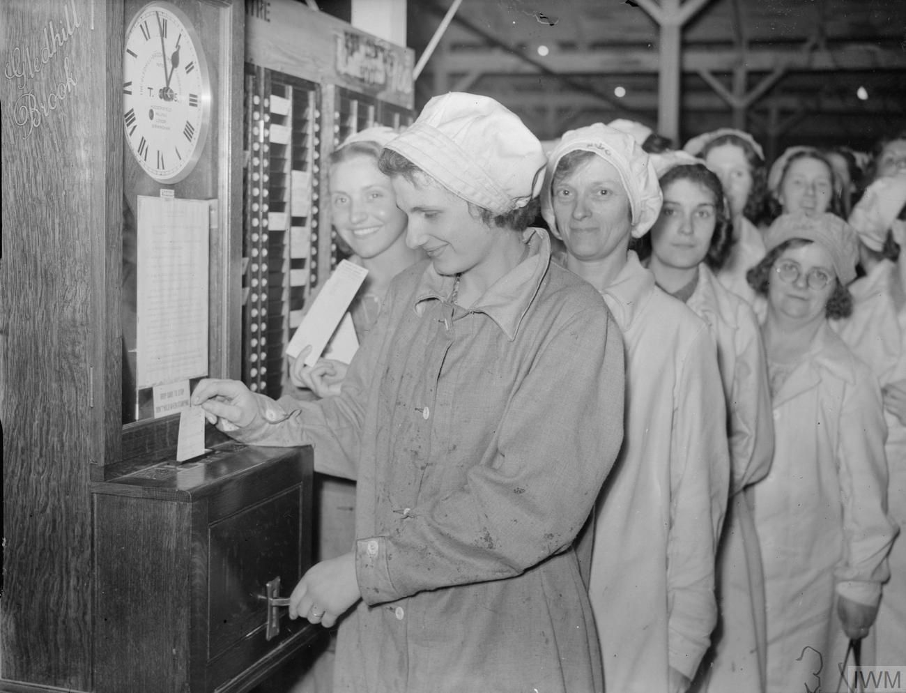 A woman in a cap pushes her time card into a factory time clock while a line of colleagues waits behind her, 1940