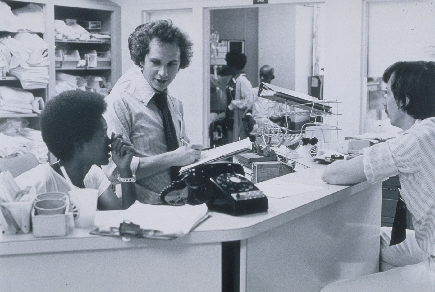 Black and white photograph of staff talking across a hospital nurses’ station with a telephone and charts